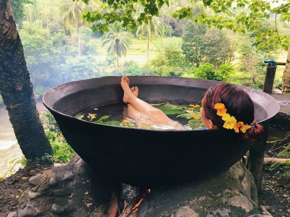 Woman relaxing in a traditional outdoor herbal bath heated over fire, surrounded by lush tropical forest and greenery in the Philippines.